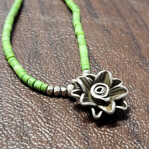 K&ouml;nnte beinhalten: Eine gr&uuml;ne Perlenkette mit einem silbernen Blumenanh&auml;nger. Die Blume ist aus gedrehtem Metall gefertigt und hat eine Spirale in der Mitte.