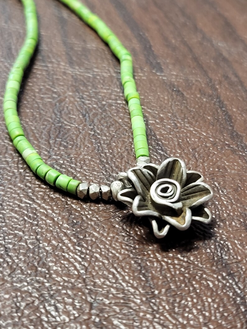 K&ouml;nnte beinhalten: Eine gr&uuml;ne Perlenkette mit einem silbernen Blumenanh&auml;nger. Die Blume ist aus gedrehtem Metall gefertigt und hat eine Spirale in der Mitte.