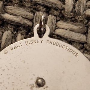 May include: A circular silver-toned metal pendant featuring a small jump ring at the top. The surface is engraved with the text WALT DISNEY PRODUCTIONS along the upper edge, accompanied by a copyright symbol. A small metal rivet is visible below the text.