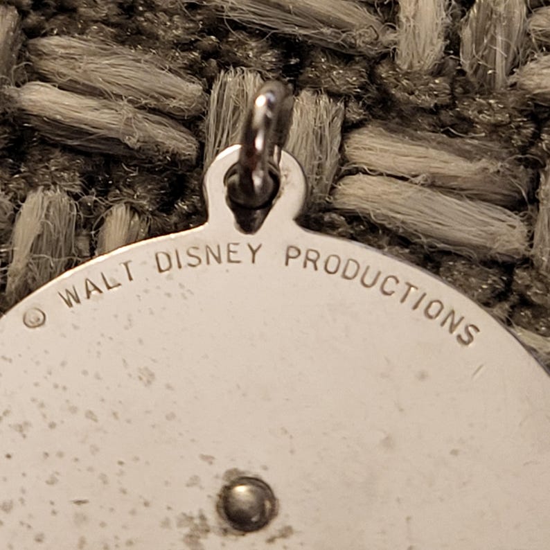 May include: A circular silver-toned metal pendant featuring a small jump ring at the top. The surface is engraved with the text WALT DISNEY PRODUCTIONS along the upper edge, accompanied by a copyright symbol. A small metal rivet is visible below the text.