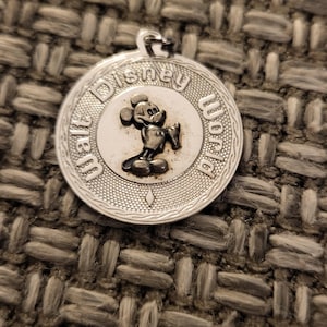 May include: A circular silver-toned metal pendant featuring a raised relief of Mickey Mouse in the center. The text Walt Disney World is embossed along the upper and right border. The outer edge has a textured pattern with a small diamond shape at the bottom.