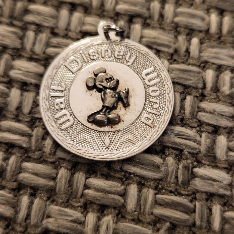 May include: A circular silver-toned metal pendant featuring a raised relief of Mickey Mouse in the center. The text Walt Disney World is embossed along the upper and right border. The outer edge has a textured pattern with a small diamond shape at the bottom.