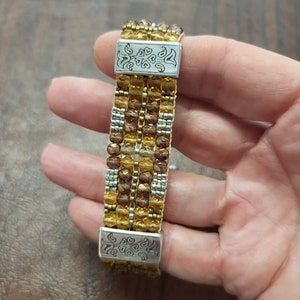 May include: A multi-strand beaded bracelet with gold and brown beads and silver metal clasps. The bracelet is made of multiple rows of beads and has a decorative design on the clasps.