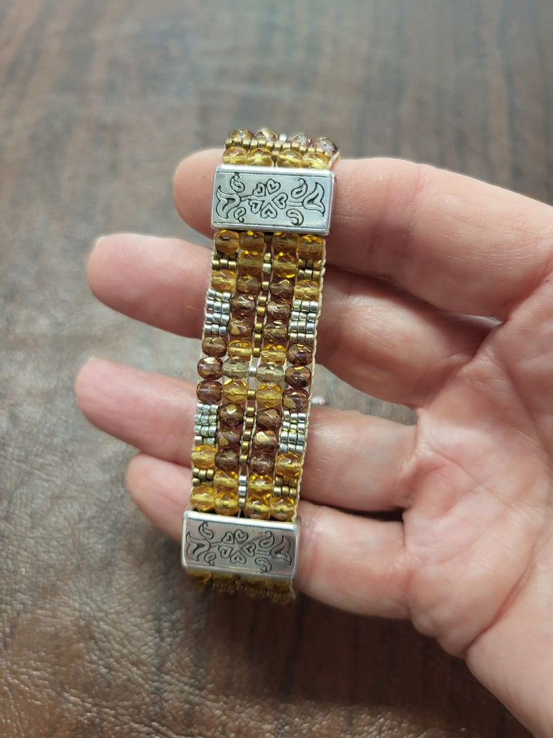 May include: A multi-strand beaded bracelet with gold and brown beads and silver metal clasps. The bracelet is made of multiple rows of beads and has a decorative design on the clasps.