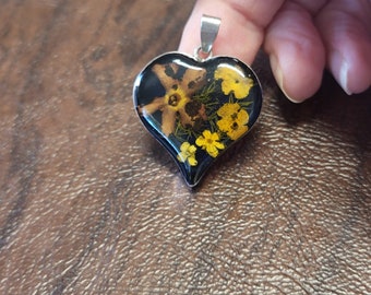 May include: Heart-shaped pendant with a silver bail, filled with dried yellow flowers and a larger brown flower against a black background. The pendant is held up by a hand.