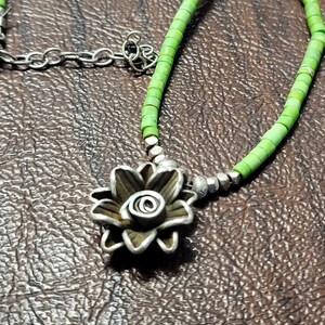 K&ouml;nnte beinhalten: Eine silberne Blumenkette mit einer gr&uuml;nen Perlenkette. Der Blumenanh&auml;nger hat ein detailliertes Design und eine Spirale in der Mitte.