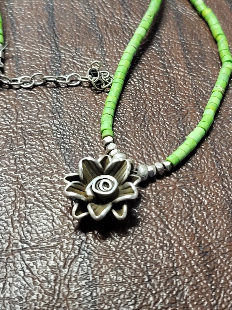 K&ouml;nnte beinhalten: Eine silberne Blumenkette mit einer gr&uuml;nen Perlenkette. Der Blumenanh&auml;nger hat ein detailliertes Design und eine Spirale in der Mitte.