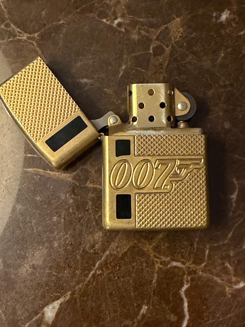 K&ouml;nnte beinhalten: Ein goldfarbenes Metallfeuerzeug mit ge&ouml;ffnetem Deckel, das ein strukturiertes Kreuzschraffurmuster und den gepr&auml;gten Text 007 auf der Vorderseite aufweist. Das Design umfasst schwarze rechteckige Akzente am Geh&auml;use und Deckel sowie ein sichtbares Z&uuml;ndrad.