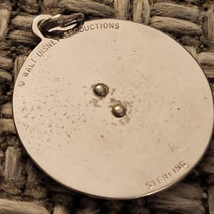 May include: A circular silver pendant featuring a small jump ring at the top. The surface is engraved with the text WALT DISNEY PRODUCTIONS along the upper edge and the word STERLING at the bottom. Two small metal rivets are visible in the center.