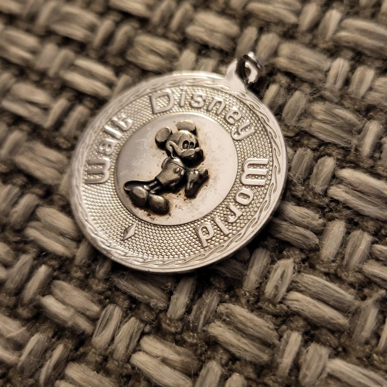 May include: A round silver-toned metal pendant featuring a raised relief of Mickey Mouse in the center. The text Walt Disney World is embossed along the outer circular border, which has a textured, cross-hatched finish and a small bail at the top.