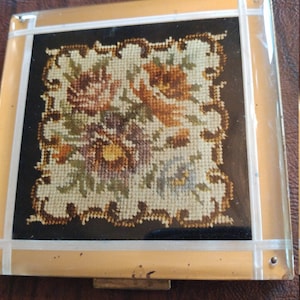 May include: A vintage gold-toned compact with a clear plastic lid. The compact features a floral design embroidered in brown, green, and white on a black background.
