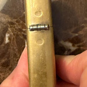 K&ouml;nnte beinhalten: Ein rechteckiges Messingfeuerzeug mit einer verwitterten, mattgoldenen Oberfl&auml;che. Das Feuerzeug verf&uuml;gt &uuml;ber ein markantes silberfarbenes Metallscharnier, das den Deckel mit dem Geh&auml;use verbindet, sowie zwei kleine kreisf&ouml;rmige Vertiefungen.