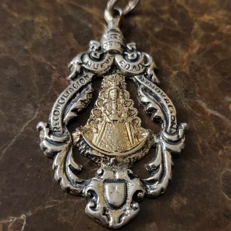 Op de afbeelding: Zilveren hanger met een gouden afbeelding van de Maagd Maria. De hanger heeft ingewikkelde details en de woorden "Reconciliacion Nueva Edad Renovacion".