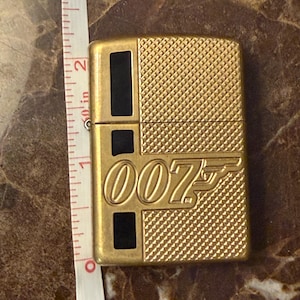 K&ouml;nnte beinhalten: Ein goldfarbenes Metallfeuerzeug mit strukturiertem Rautenmuster und schwarzen rechteckigen Akzenten. Auf der Vorderseite befindet sich das 007-Logo in erhabener Schrift. Ein wei&szlig;es Ma&szlig;band daneben zeigt eine H&ouml;he von etwa 5,7 cm.
