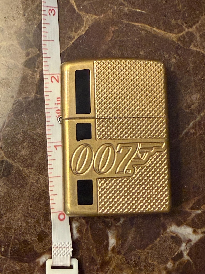 K&ouml;nnte beinhalten: Ein goldfarbenes Metallfeuerzeug mit strukturiertem Rautenmuster und schwarzen rechteckigen Akzenten. Auf der Vorderseite befindet sich das 007-Logo in erhabener Schrift. Ein wei&szlig;es Ma&szlig;band daneben zeigt eine H&ouml;he von etwa 5,7 cm.
