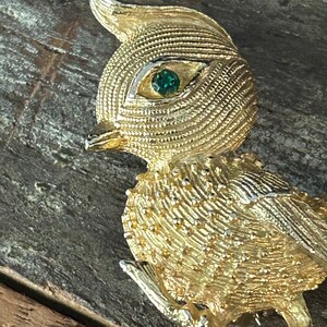 May include: A gold-toned bird brooch with a green rhinestone eye. The bird is perched on a branch and has a textured surface.