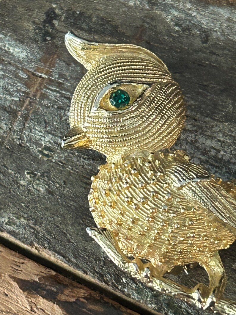 May include: A gold-toned bird brooch with a green rhinestone eye. The bird is perched on a branch and has a textured surface.