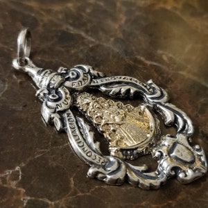 Op de afbeelding: Zilveren hanger met een goudkleurig beeld van de Maagd Maria. De hanger heeft ingewikkelde details en de inscriptie "RECONCILIACION" aan de onderkant.