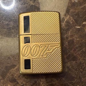 K&ouml;nnte beinhalten: Ein goldfarbenes Feuerzeug aus Metall mit einem strukturierten Rautenmuster und drei schwarzen rechteckigen Einlagen auf der linken Seite. Die Vorderseite zeigt das gepr&auml;gte 007-Logo mit dem ikonischen Pistolenlauf-Design in der Ziffer sieben.