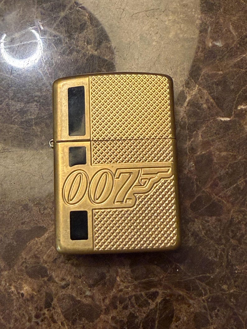 K&ouml;nnte beinhalten: Ein goldfarbenes Feuerzeug aus Metall mit einem strukturierten Rautenmuster und drei schwarzen rechteckigen Einlagen auf der linken Seite. Die Vorderseite zeigt das gepr&auml;gte 007-Logo mit dem ikonischen Pistolenlauf-Design in der Ziffer sieben.