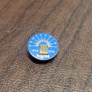 May include: A round, blue and gold Centennial pin with the text "Centennial" around the edge. The center features a sunburst design above a building, with the years 1858 and 1958 on either side. The word "SQUIBB" is printed below.