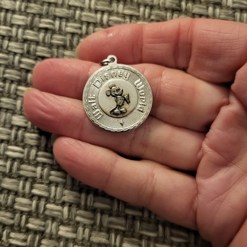 May include: A circular silver-toned metal charm featuring a raised relief of Mickey Mouse in the center. The text Walt Disney World is embossed along the border of the charm, which has a textured outer rim and a small loop at the top for attachment.