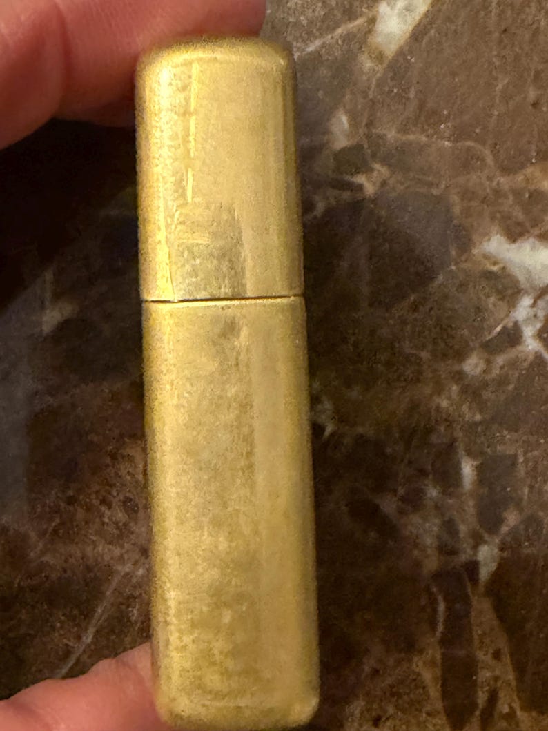 K&ouml;nnte beinhalten: Ein rechteckiges, goldfarbenes Feuerzeug aus Metall mit geb&uuml;rsteter Oberfl&auml;che und Klappdeckel. Das Feuerzeug hat eine vertikale Ausrichtung mit abgerundeten Ecken und eine glatte, metallische Oberfl&auml;chenstruktur, die das Licht reflektiert.