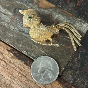 May include: Gold-tone bird brooch with green rhinestone eyes and a long, braided tail. The brooch is about the size of a US quarter.