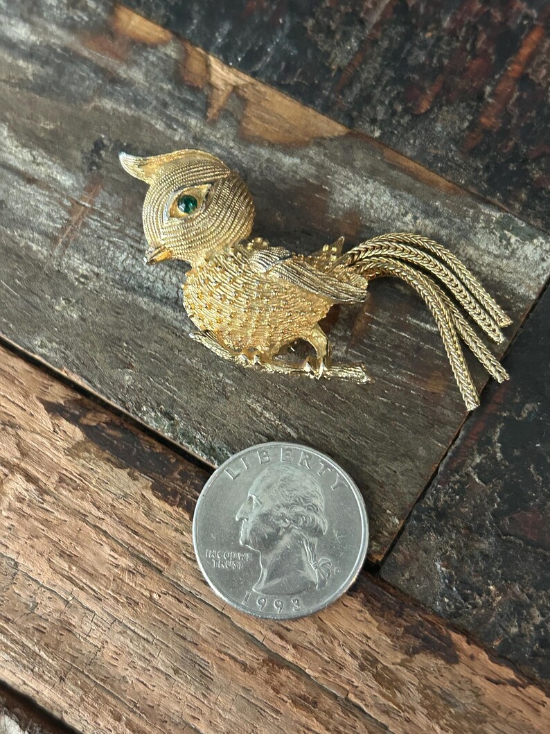 May include: Gold-tone bird brooch with green rhinestone eyes and a long, braided tail. The brooch is about the size of a US quarter.