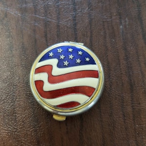 May include: A round, silver compact with a hinged lid. The lid features a red, white, and blue American flag design.