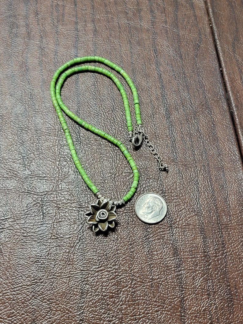 K&ouml;nnte beinhalten: Eine gr&uuml;ne Perlenkette mit einem silbernen Blumenanh&auml;nger. Die Kette ist etwa 45 cm lang und der Anh&auml;nger etwa 2,5 cm im Durchmesser.