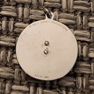 May include: A circular silver pendant featuring a small metal loop at the top. The surface is engraved with the text WALT DISNEY PRODUCTIONS at the top and STERLING at the bottom. Two small metal rivets are positioned in the center of the pendant.