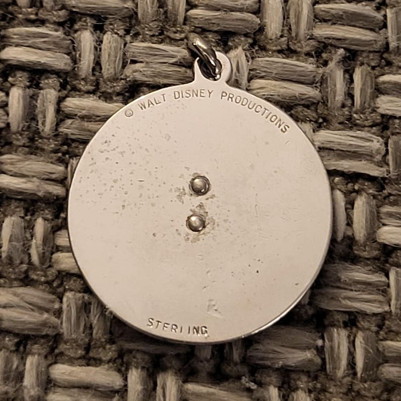 May include: A circular silver pendant featuring a small metal loop at the top. The surface is engraved with the text WALT DISNEY PRODUCTIONS at the top and STERLING at the bottom. Two small metal rivets are positioned in the center of the pendant.