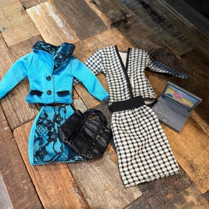 May include: Miniature doll clothing set featuring a light blue jacket and skirt with black lace accents, a black and white checkered jacket and skirt, a black handbag, and a small laptop. The doll clothes are arranged on a wooden surface.