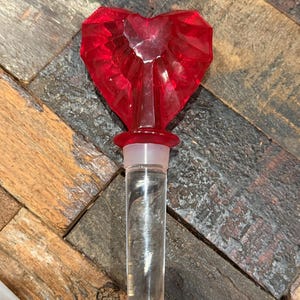 May include: A bottle stopper featuring a faceted, translucent red heart-shaped top attached to a clear, tapered cylindrical base. The stopper includes a translucent silicone ring at the junction between the red heart and the clear bottom section.
