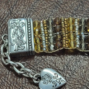 May include: A silver bracelet with a heart charm and a clasp featuring a floral design. The bracelet is made of beads in shades of brown, gold, and silver.