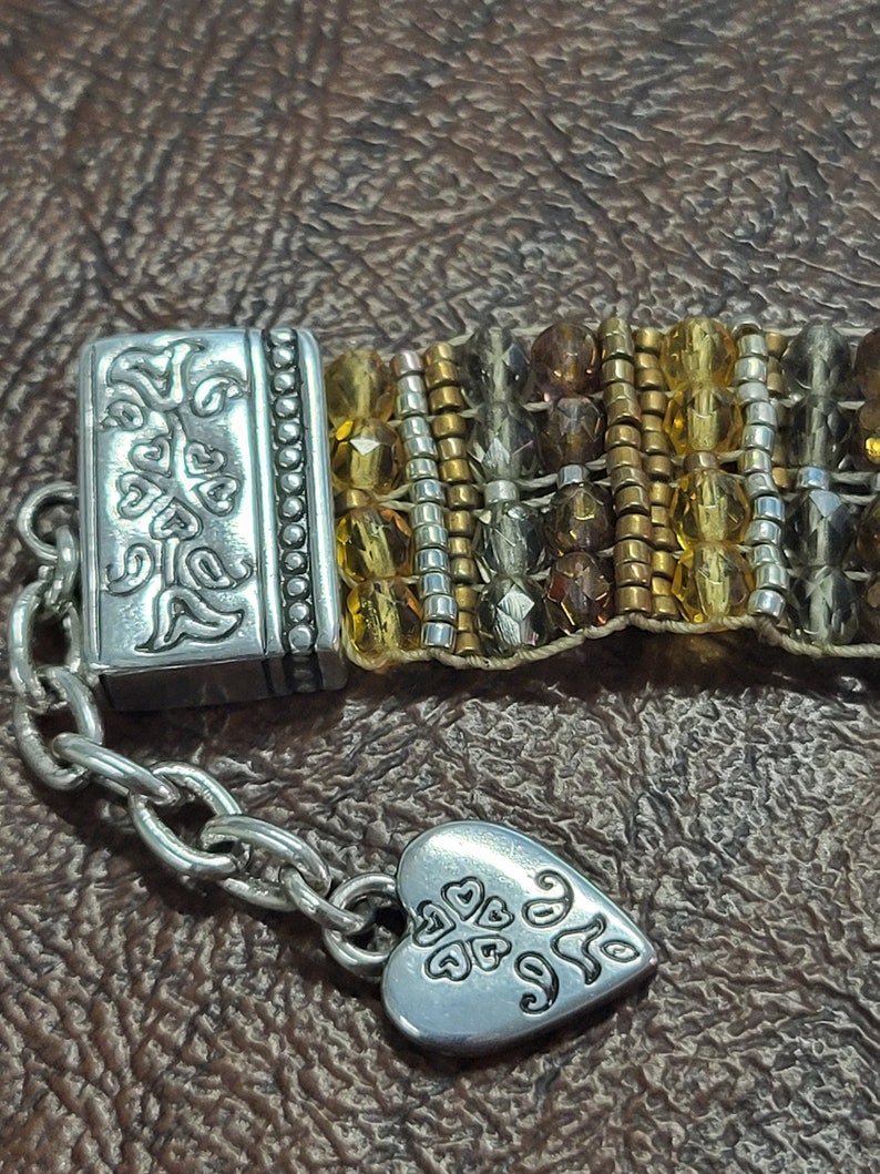 May include: A silver bracelet with a heart charm and a clasp featuring a floral design. The bracelet is made of beads in shades of brown, gold, and silver.