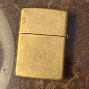 K&ouml;nnte beinhalten: Ein rechteckiges Messingfeuerzeug mit Klappdeckel und einer verwitterten, matten Metalloberfl&auml;che. Das Feuerzeug hat ein schlichtes, klares Design mit abgerundeten Ecken und einer sichtbaren horizontalen Naht, die den Deckel vom Hauptk&ouml;rper trennt.