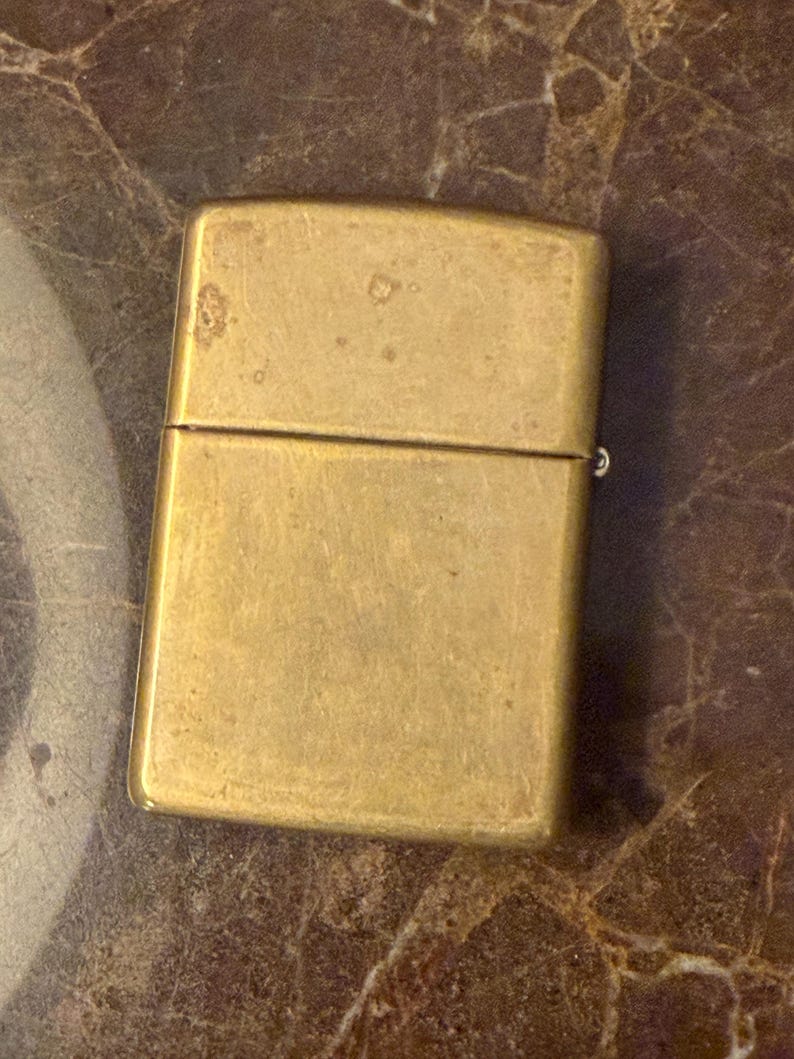 K&ouml;nnte beinhalten: Ein rechteckiges Messingfeuerzeug mit Klappdeckel und einer verwitterten, matten Metalloberfl&auml;che. Das Feuerzeug hat ein schlichtes, klares Design mit abgerundeten Ecken und einer sichtbaren horizontalen Naht, die den Deckel vom Hauptk&ouml;rper trennt.