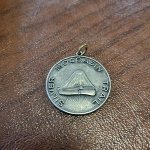 May include: A round, silver-toned metal pendant featuring an embossed design of a mountain and a pair of moccasins. The text 'SILVER MOCCASIN TRAIL' is engraved along the circular border. A small metal jump ring is attached to the top of the pendant.