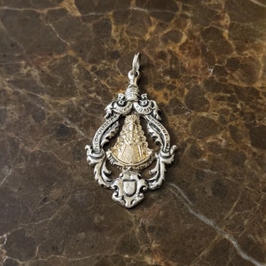 Op de afbeelding: Zilveren hanger met een gouden afbeelding van de Maagd Maria. De hanger heeft de tekst "NUEVA EDAD" en "RECONCILIACION" boven en onder de afbeelding.