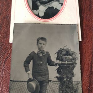 May include: A black and white photo of two children. The younger child is wearing a hat and a dress. The older child is wearing a suit and holding a hat. The older child is standing next to a potted plant.