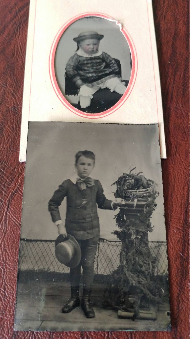 May include: A black and white photo of two children. The younger child is wearing a hat and a dress. The older child is wearing a suit and holding a hat. The older child is standing next to a potted plant.