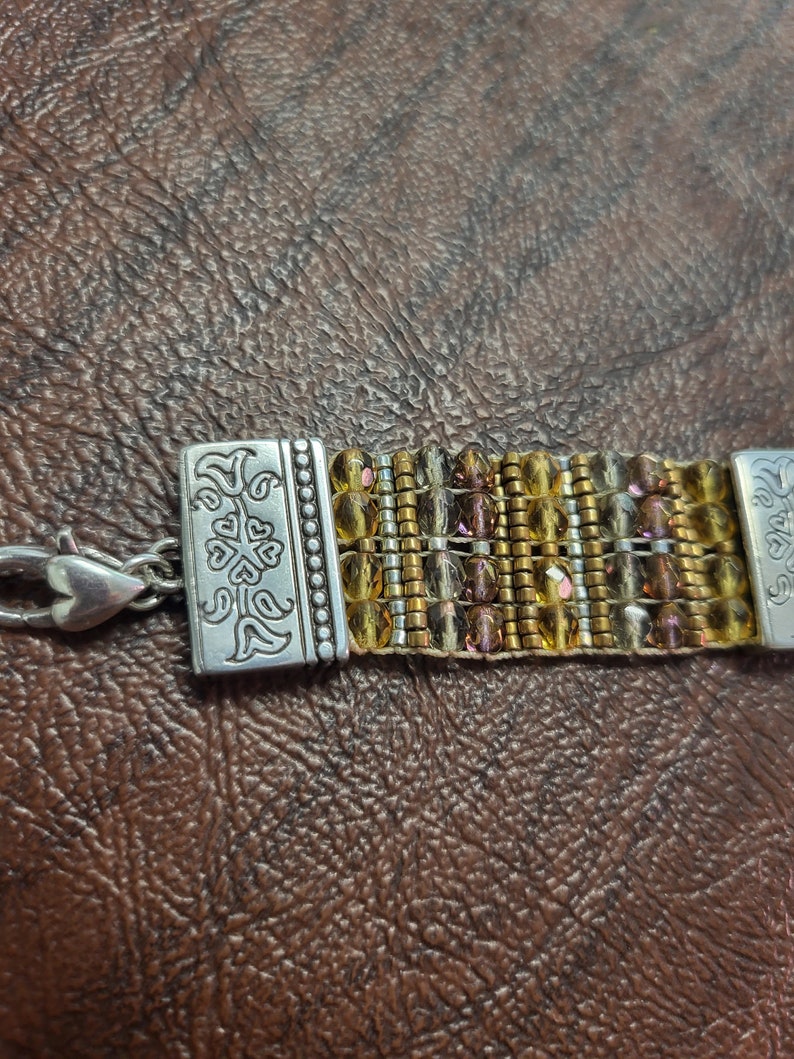 May include: A gold and brown beaded bracelet with a silver clasp. The clasp has a floral design.