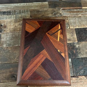 May include: A wooden box with a geometric pattern made of different types of wood. The box has a dark brown frame and a top made of various shades of brown, red, and yellow wood.