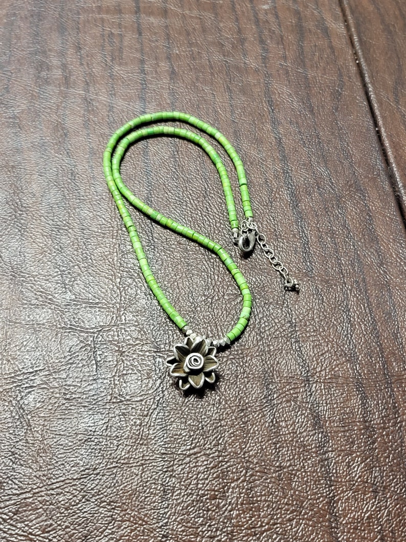 K&ouml;nnte beinhalten: Eine gr&uuml;ne Perlenkette mit einem silbernen Blumenanh&auml;nger. Die Kette besteht aus kleinen, runden Perlen und hat ein schlichtes, elegantes Design.