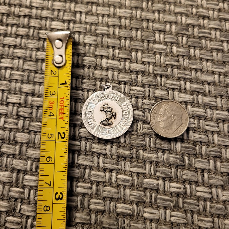 May include: A round silver-toned metal charm featuring a raised Mickey Mouse figure and the text Walt Disney World. The charm is positioned next to a yellow measuring tape showing a diameter of approximately 1.25 inches and a US dime coin.