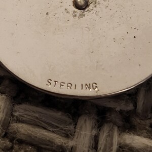 May include: A circular silver object with a smooth, metallic surface featuring a small central protrusion. The word STERLING is clearly stamped in capital letters along the bottom edge of the curved surface, indicating the metal composition.