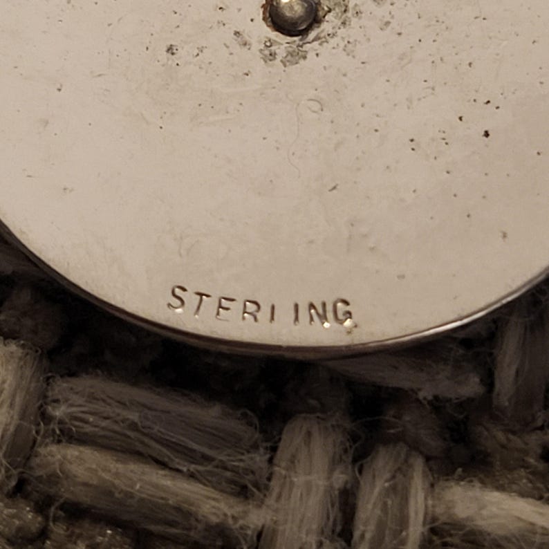 May include: A circular silver object with a smooth, metallic surface featuring a small central protrusion. The word STERLING is clearly stamped in capital letters along the bottom edge of the curved surface, indicating the metal composition.