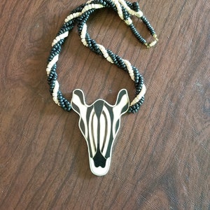May include: A zebra-themed necklace featuring a pendant shaped like a zebra's head. The pendant has black and white stripes. The necklace is made of black and white beads, and the background is a brown surface.